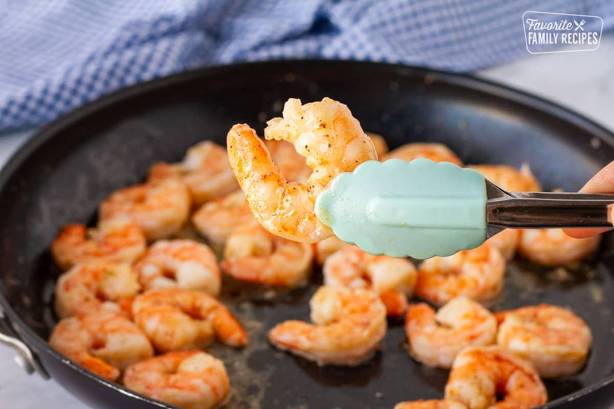 Tongs holding a cooked shrimp over the skillet of cooked shrimp.