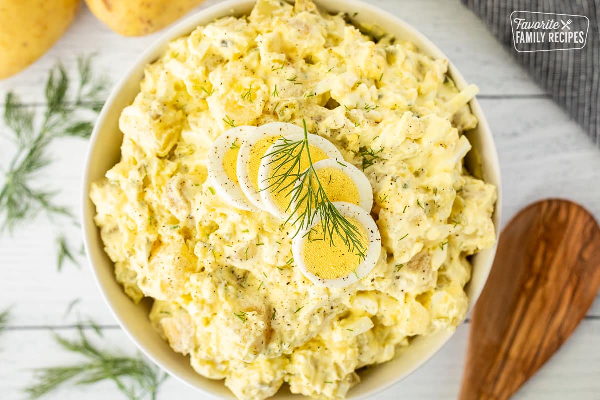 Bowl of Homemade Potato Salad with sliced hard boiled eggs on top and fresh dill. Wooden spoon on the side.