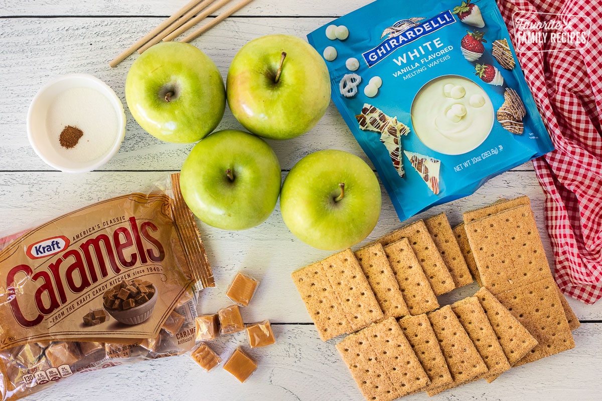 Ingredients to make Apple Pie Caramel Apples including apples, caramels, cinnamon graham crackers, melting while chocolate, green apples, wooden sticks, sugar and cinnamon.