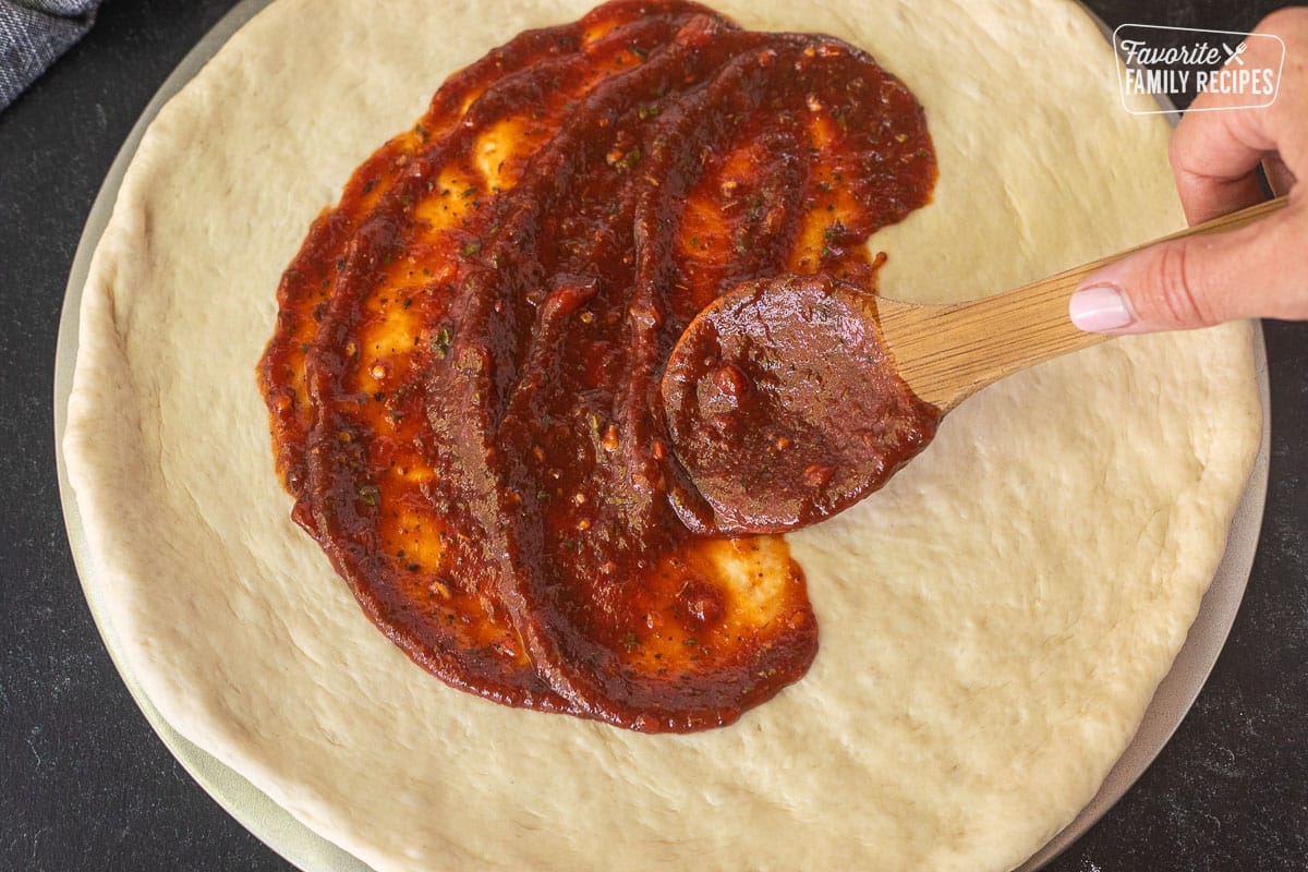 Spreading pizza sauce with a wooden spoon on a rolled out pizza dough.