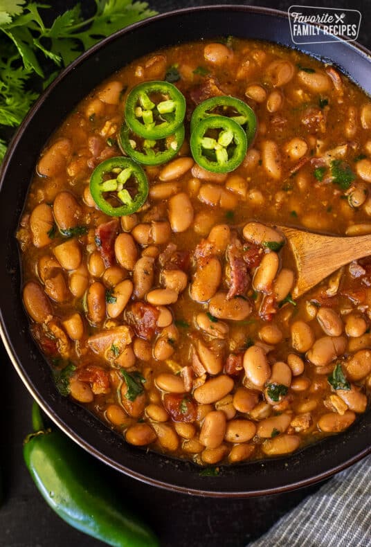 Charro Beans in a bowl with a wooden spoon and garnished with sliced jalapeños.