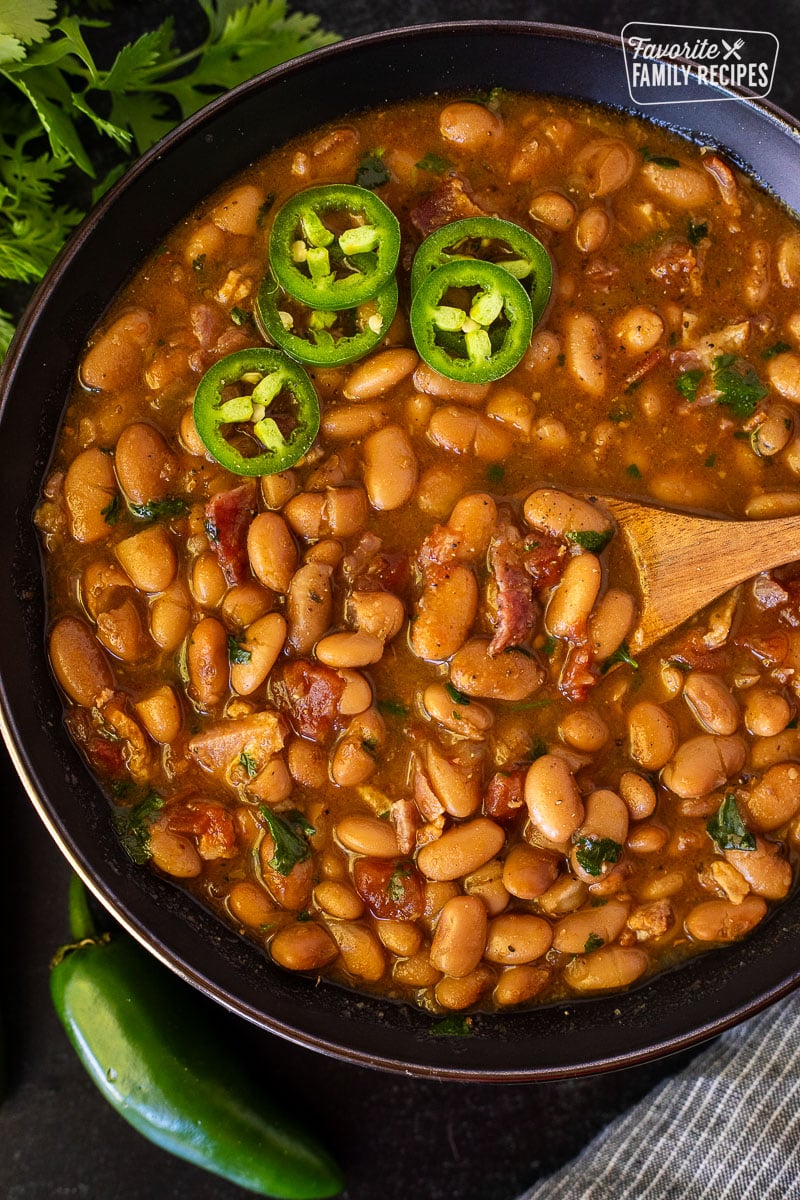 Charro Beans in a bowl with a wooden spoon and garnished with sliced jalapeños.