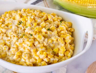 Bowl of Creamed Corn garnished with fresh parsley, salt and pepper. Fresh corn in the background.