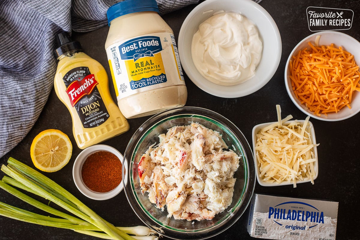 Ingredients to make Hot Crab Dip including crab, cheddar cheese, parmesan cheese, mayo, sour cream, cream cheese, dijon mustard, lemon, Old Bay seasoning and green onions.