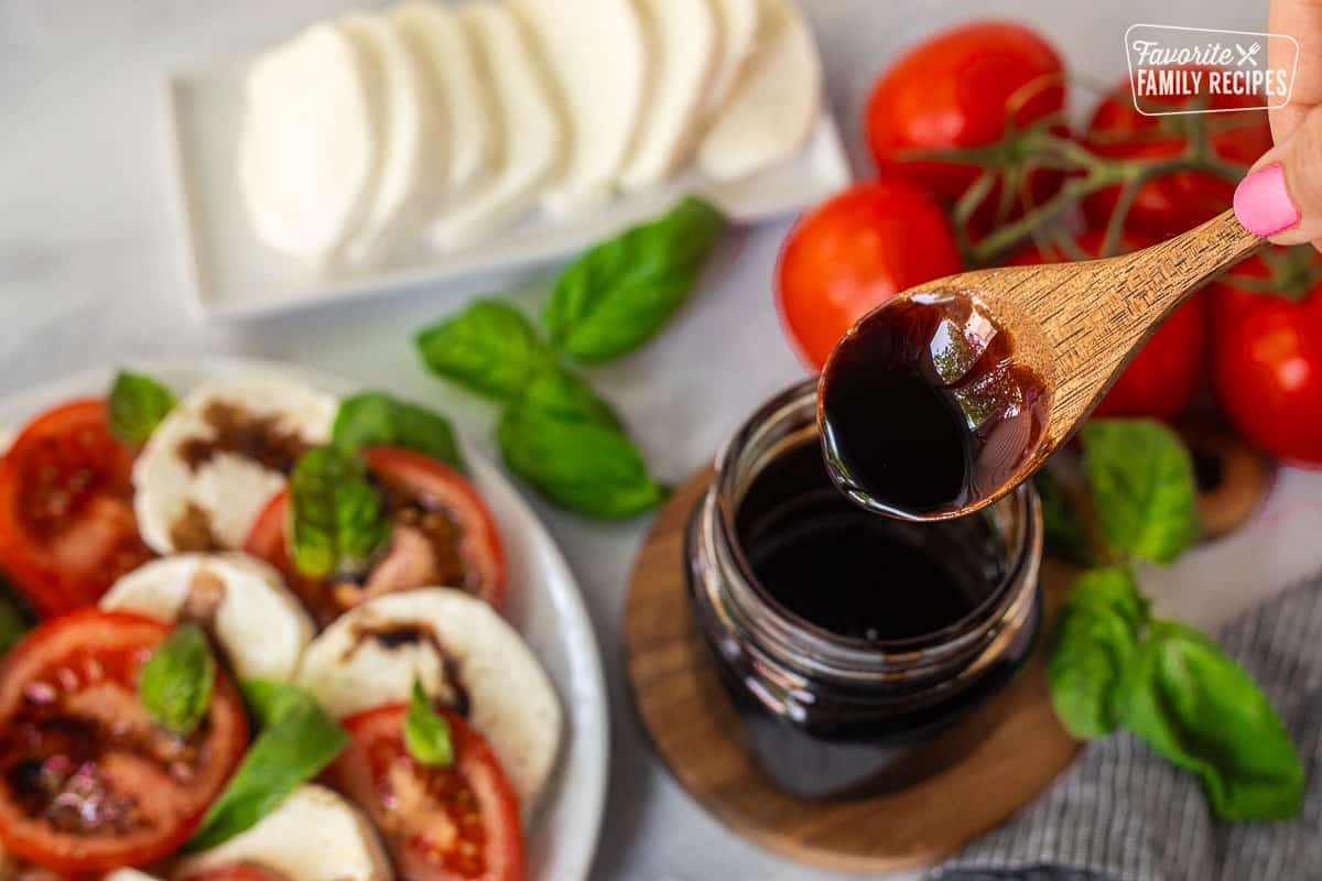 Wooden spoon of balsamic glaze over jar next to caprese salad.