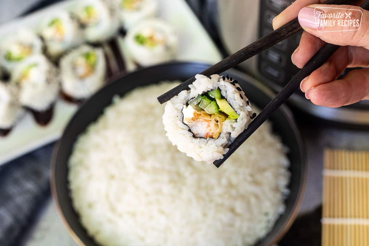 Holding a sushi roll with chopsticks in from tof a bowl of sushi rice.