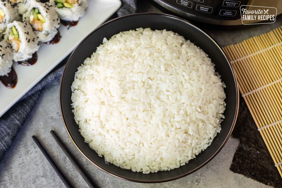 Bowl of Instant Pot Sushi Rice next to an instant pot, chop stick, tray of sushi rolls and bamboo mat.