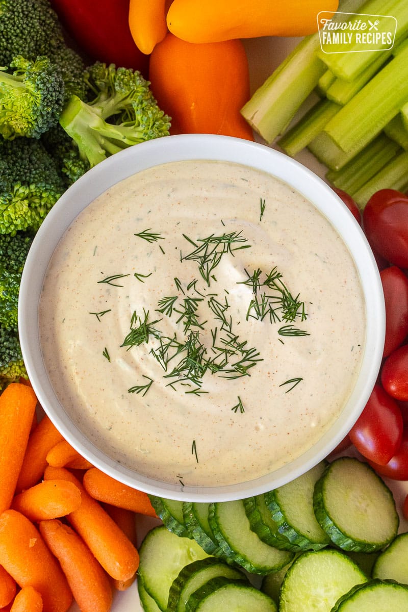 Bowl of Veggie Dip topped with dill and surrounded by fresh vegetables.