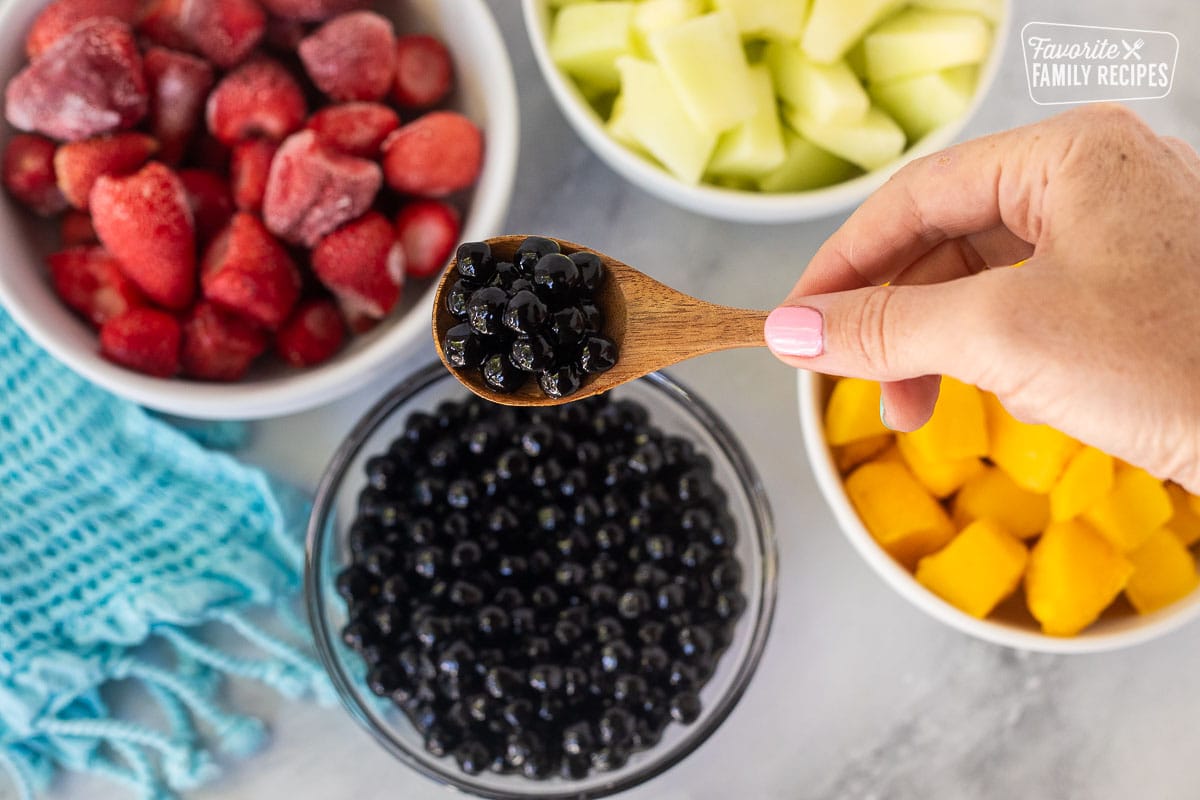 Spoon of black tapioca peals held up by a hand. Bowls of frozen strawberries, honeydew melon and mango in the background.