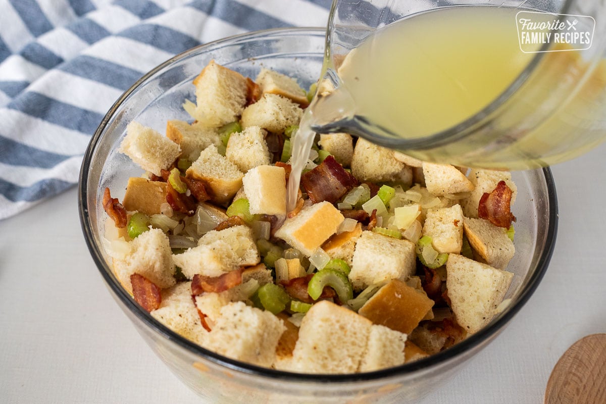 Pouring chicken broth on top of bowl of cubed bread, bacon, celery and onion mixture.