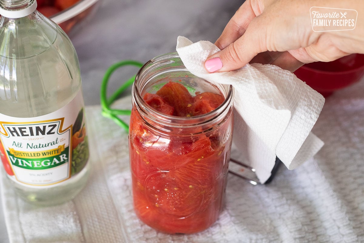 Wiping the rim of the jar with white vinegar on a paper towel. Jar of white vinegar on the side.