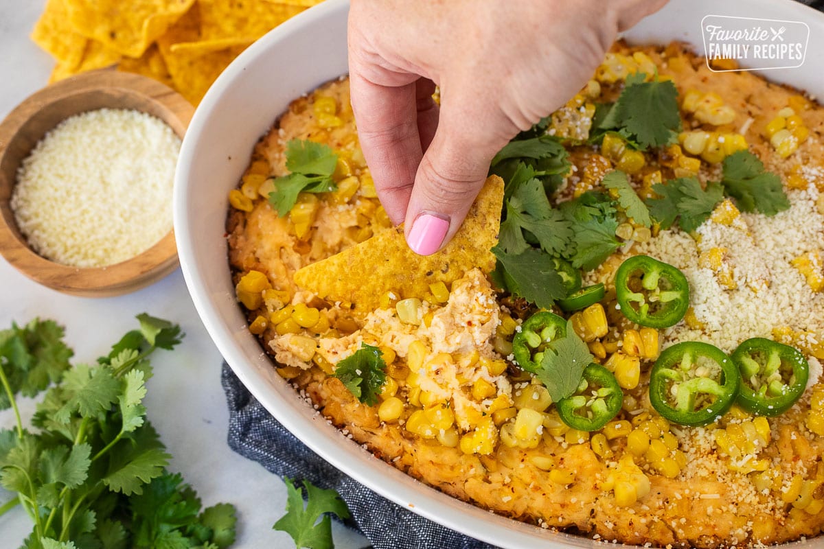 Hand dipping a tortilla chip into Mexican Street Corn Dip in a dish toped with jalapeño, cilantro and cojita cheese.