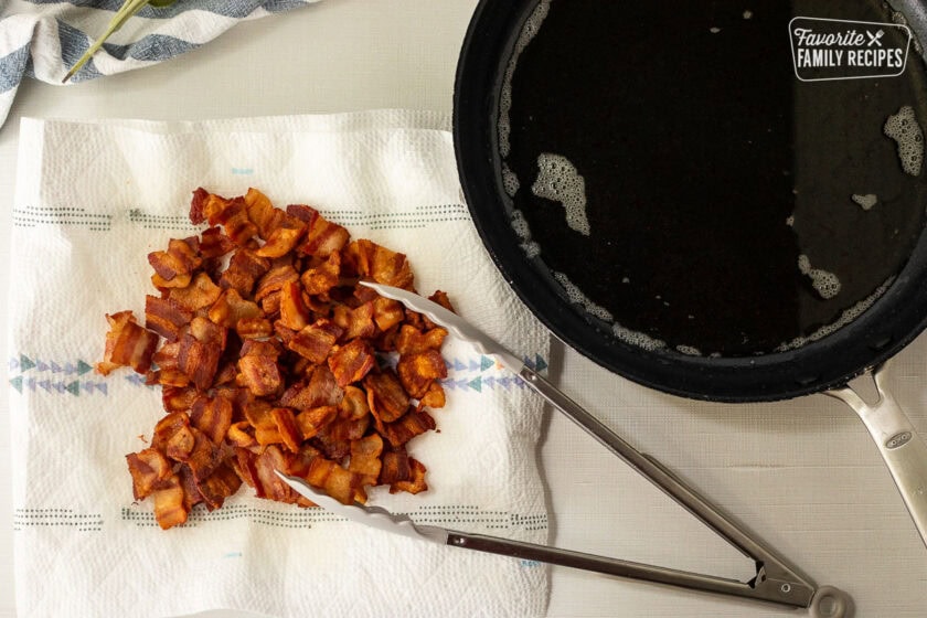 Paper towel with chopped cooked bacon with tongs next to skillet with bacon grease.