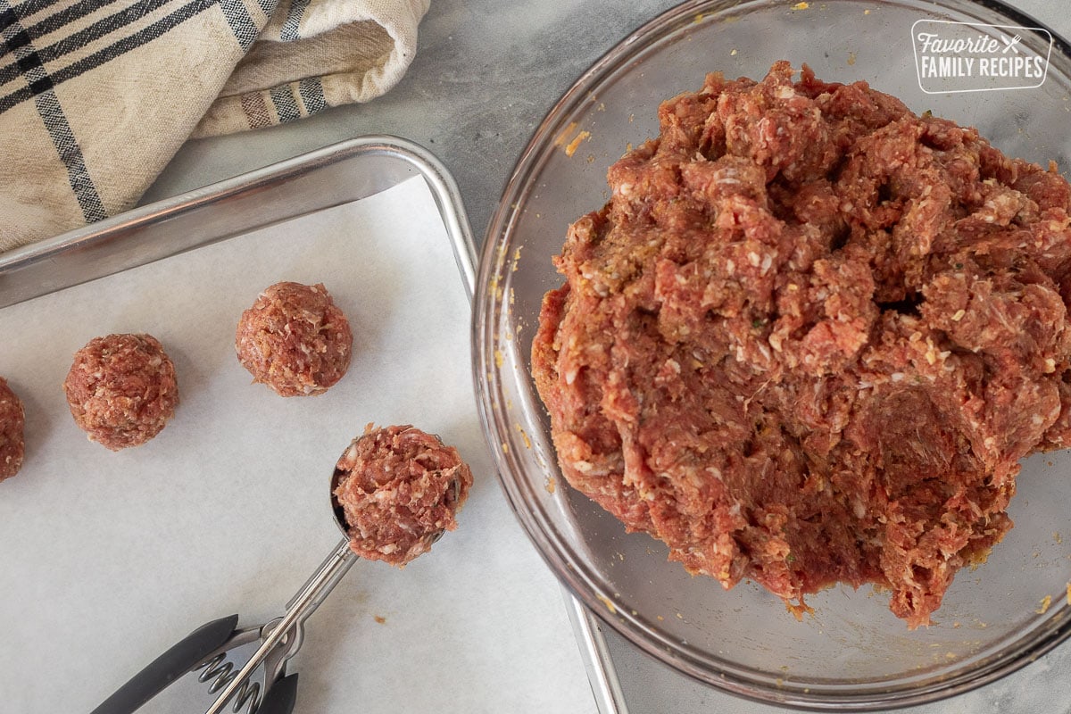 Scooped meatballs on a cookie sheet next to a bowl of meat mixture.