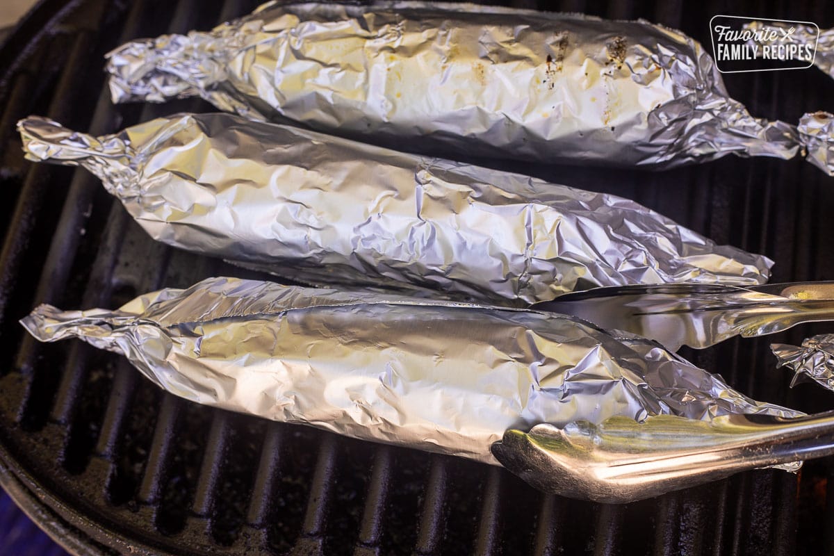 Tongs flipping grilled corn on the cob wrapped in foil.