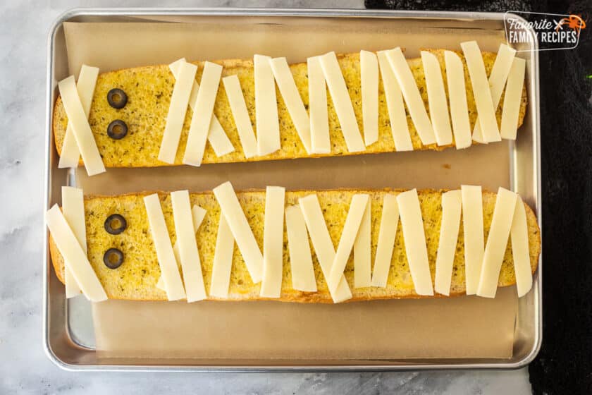 Baking sheet lined with parchment paper, garlic bread and strips of mozzarella cheese and olive slices for eyes.