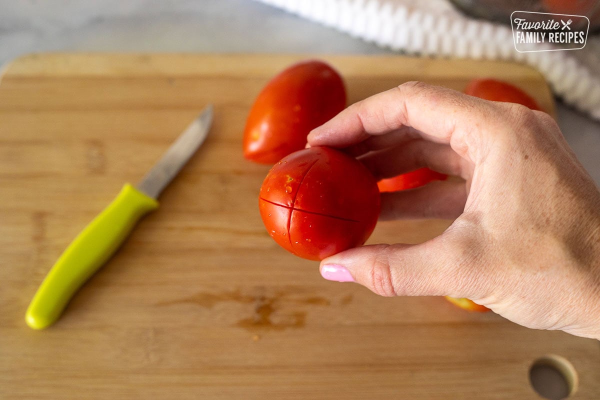 Cut lines on the bottom of a Roma tomato held over a cutting board with a knife and tomatoes. .