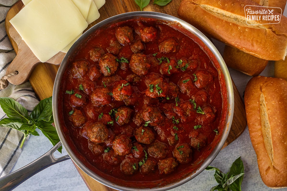 Meatballs in marinara sauce in skillet surrounded by rolls and cheese.