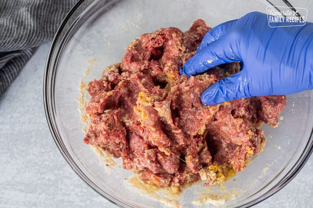 Mixing together meatballs with a gloved hand in a glass bowl.