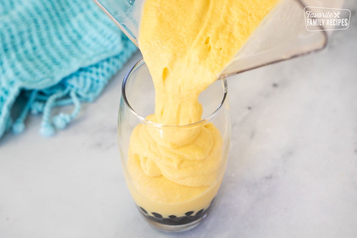 Pouring mango smoothie from a blender into a glass with black tapioca pearls on the bottom.