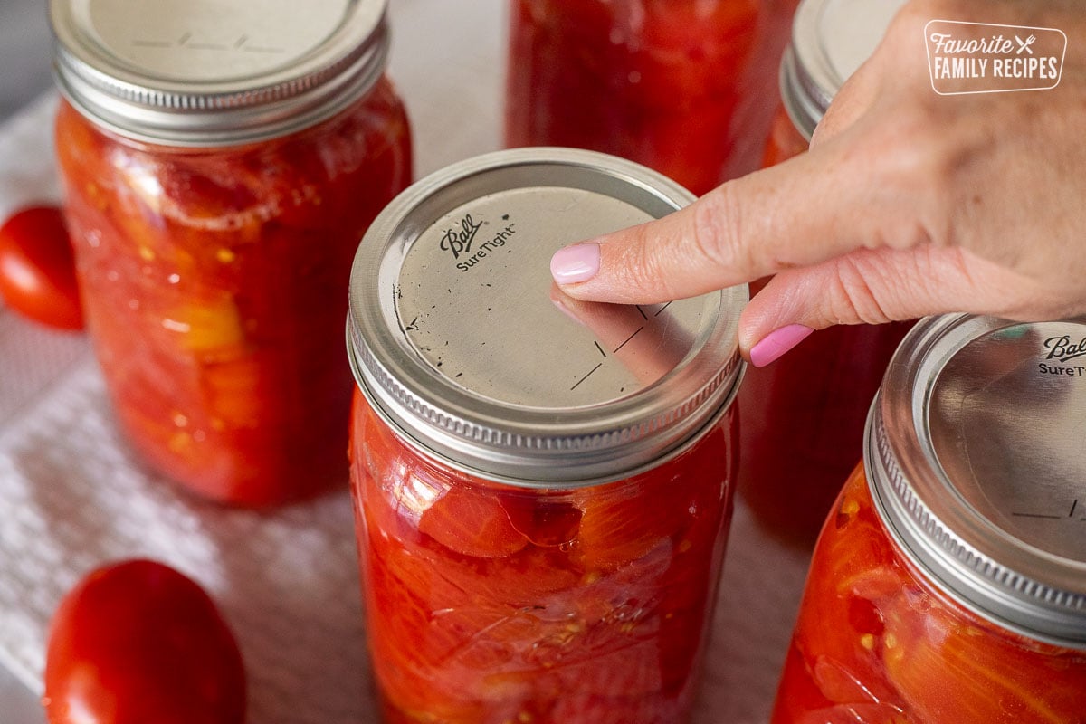 Finger pressing the top lid of a canning jar of tomatoes.