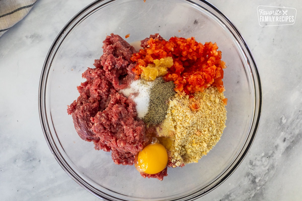 Glass mixing bowl with ground beef, egg, cracker crumbs, salt and pepper, ginger, onion, milk, and red bell peppers.