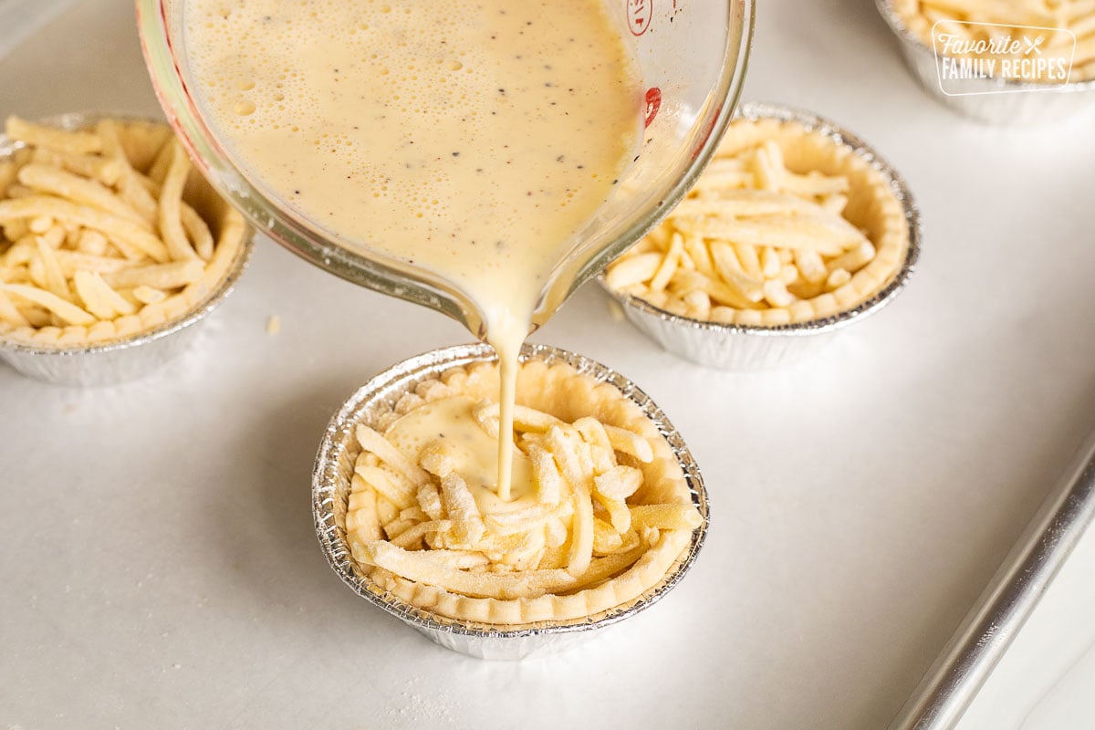 Pouring egg mixture into pie shells filled with shredded cheese.