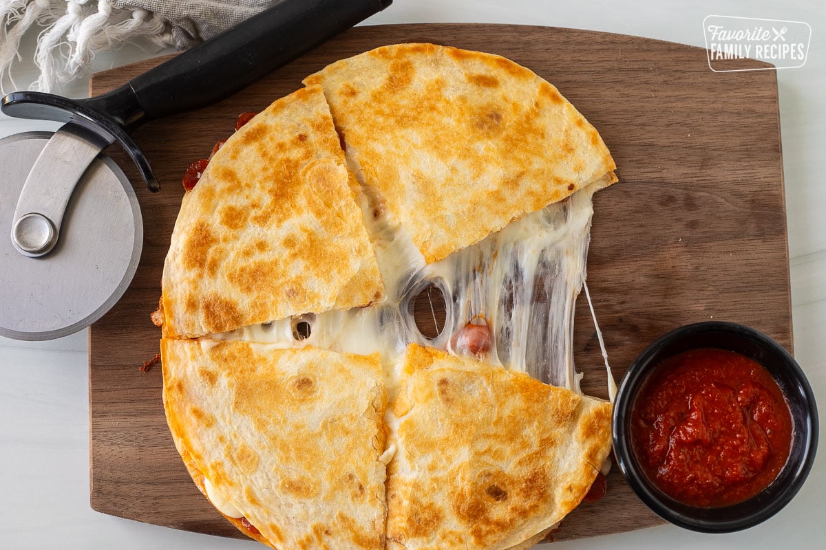 Pizza quesadilla on a cutting board with cheese stretched and a side cup of pizza sauce.