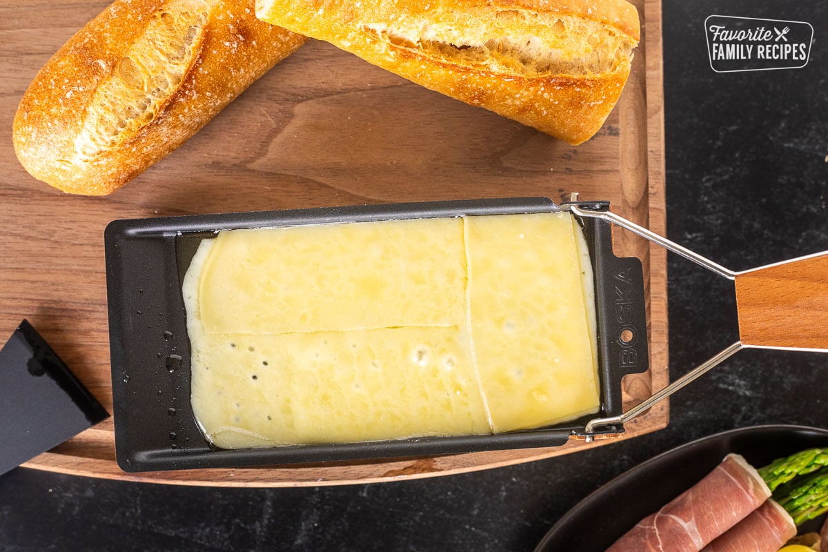 Melted cheese in a raclette dish on a cutting board next to baguettes.