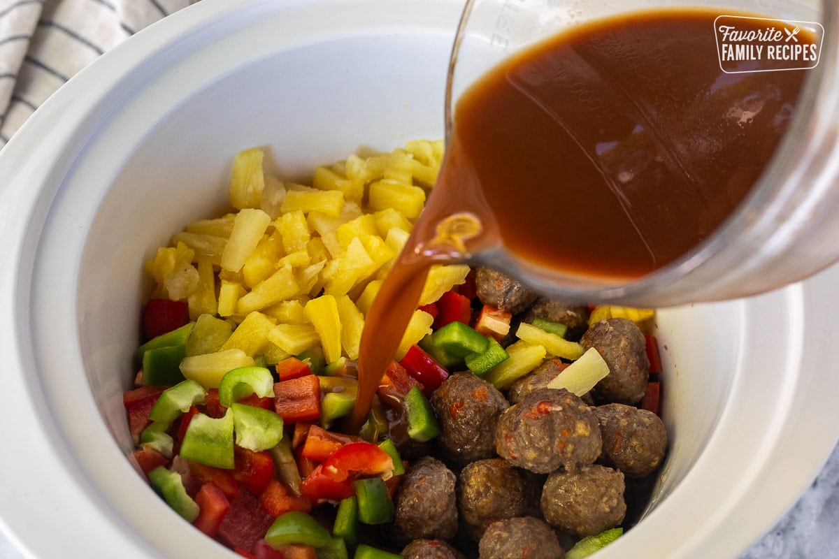 Pouring sweet and sour sauce in a crock pot of meatballs, pineapple, and bell peppers.