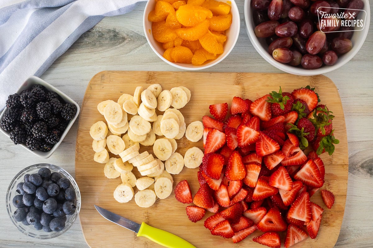 Cut up strawberries and bananas on a cutting board next to bowls of grapes, mandarin oranges, blackberries and blueberries.