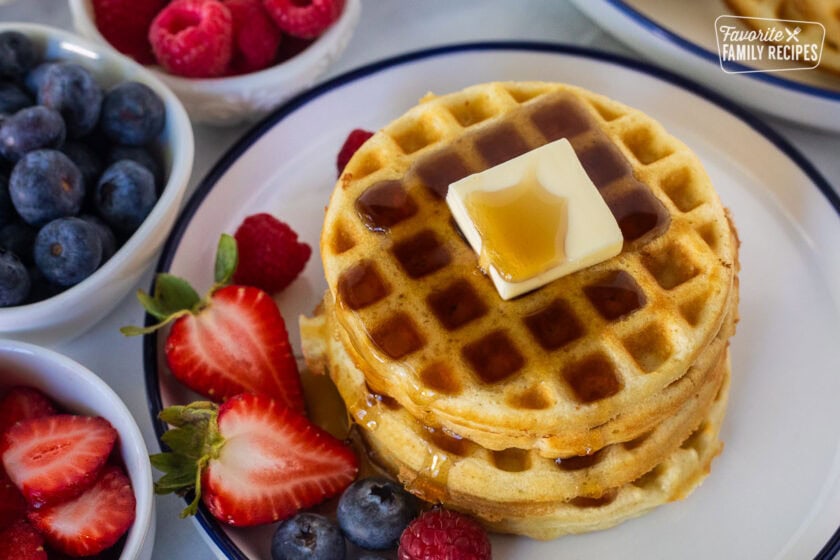 Easy homemade waffles stacked on a plate with butter and syrup. Fresh strawberries, blueberries and raspberries on the side.