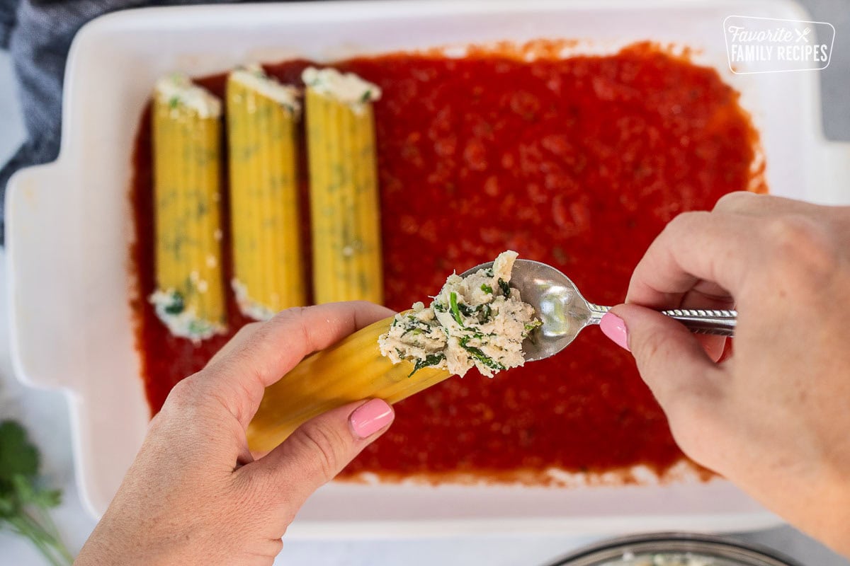 Filling dry manicotti shells with chicken and spinach filling using a spoon.