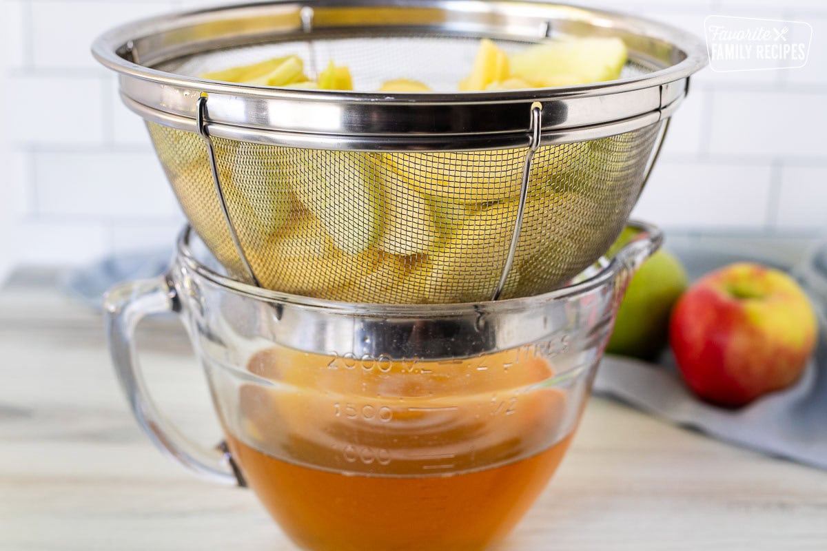 Colander with sliced apples over bowl of apple juice.