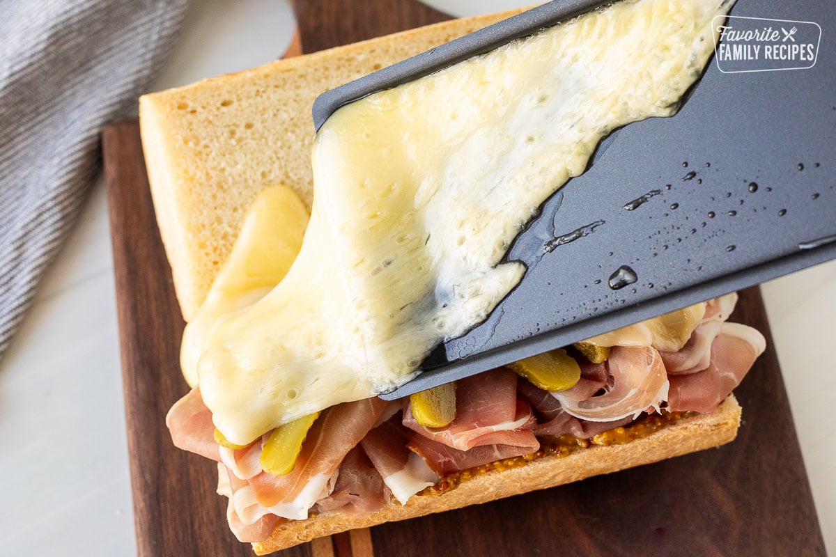 Pouring melty raclette cheese on top of a prosciutto sandwich.