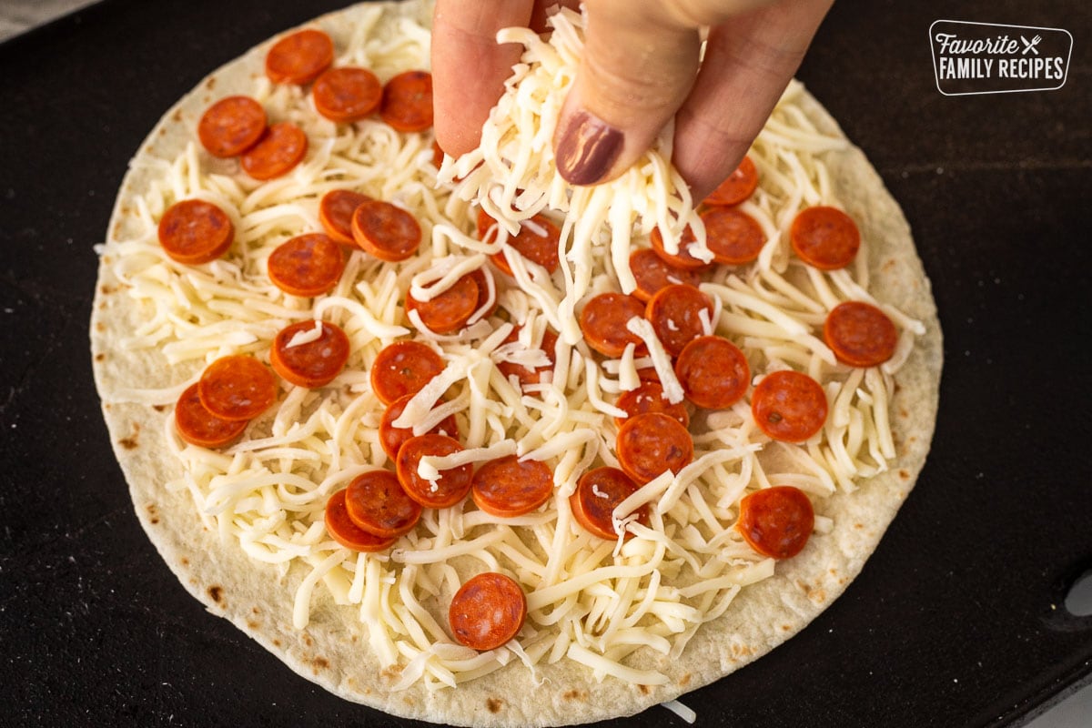 Sprinkling mozzarella cheese on top of a tortilla with shredded mozzarella cheese on a skillet with mini pepperoni.