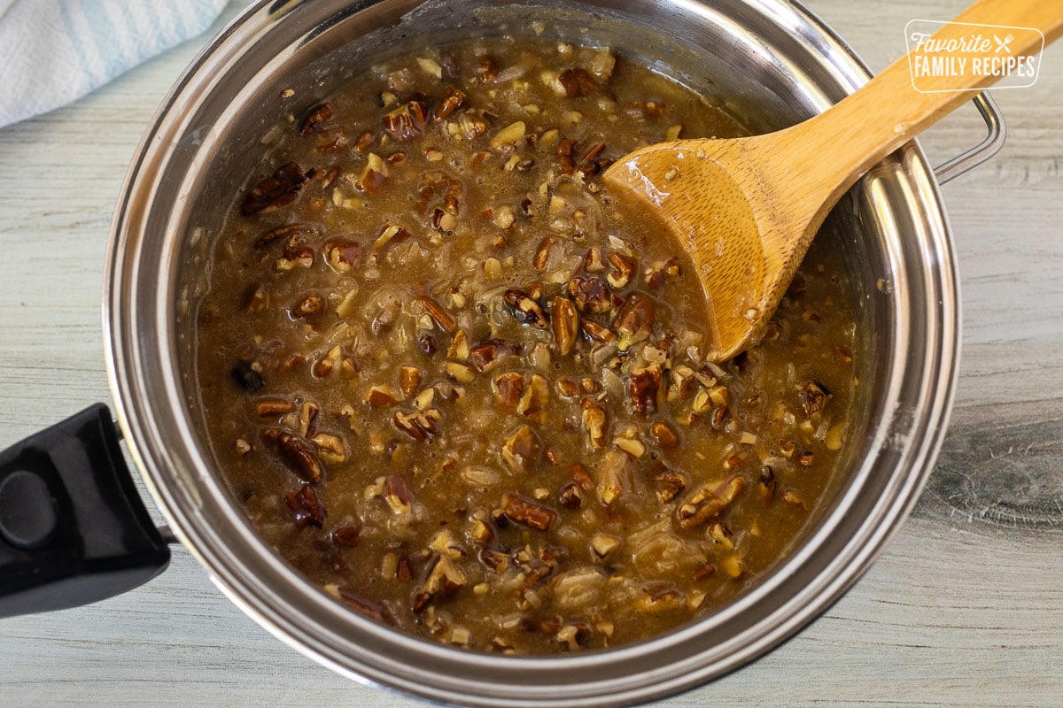 Saucepan with pecans and caramel added to brown sugar butter mixture with a wooden spoon resting in the pan.