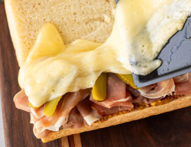 Pouring melty raclette cheese on top of a sandwich.