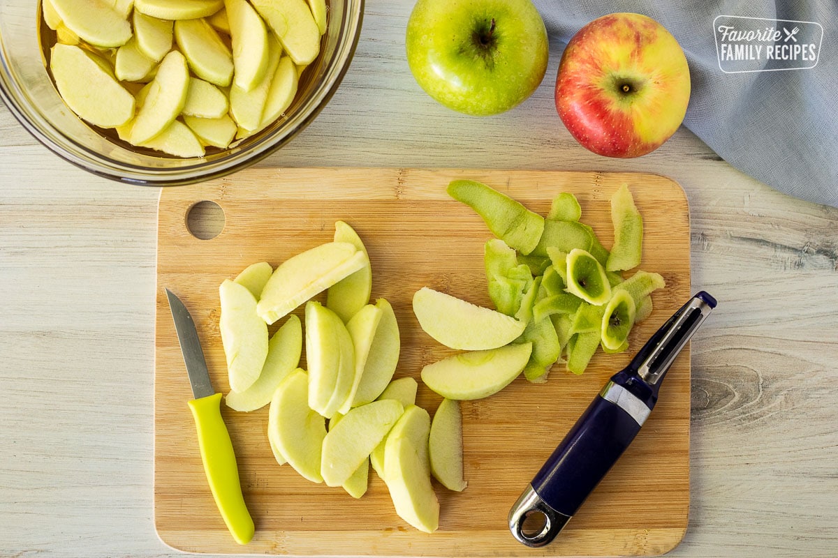 Cutting board with sliced and peeled apples next to a bowl of apples in apple juice.