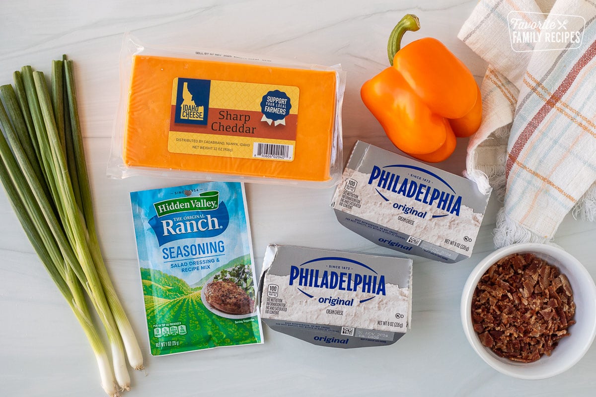 Ingredients to make a Pumpkin Shaped Cheese Ball including sharp cheddar cheese, bell pepper, cream cheese, bacon, ranch seasoning packet and green onions.