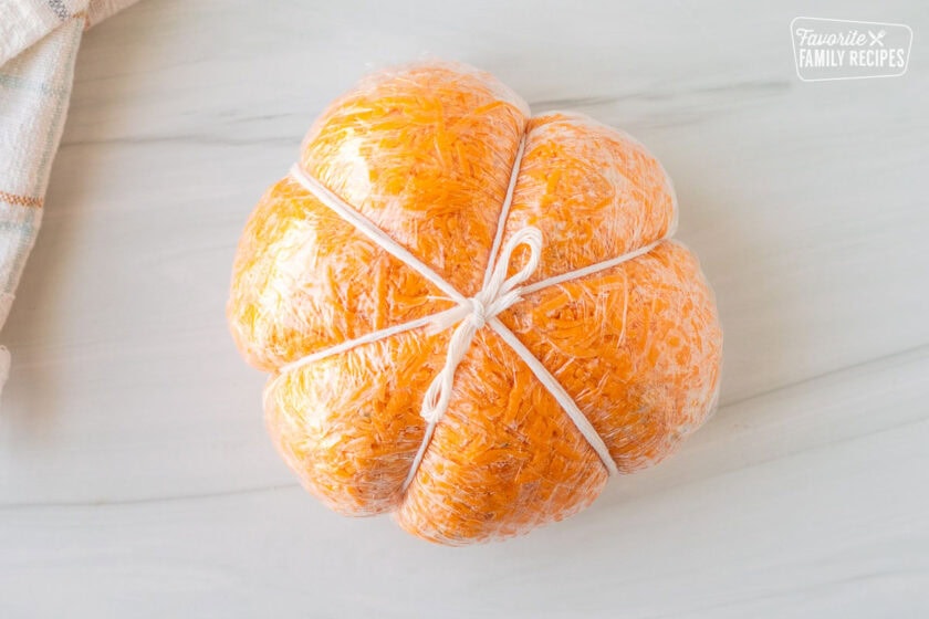 Pumpkin shaped cheese ball wrapped in plastic wrap with twine tied in a knot.