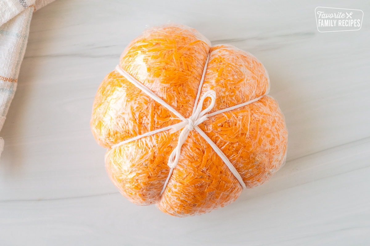 Pumpkin shaped cheese ball wrapped in plastic wrap with twine tied in a knot.