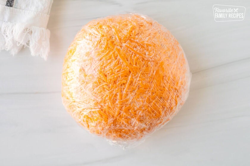 Cheese ball covered in shredded cheddar cheese and wrapped in plastic wrap.