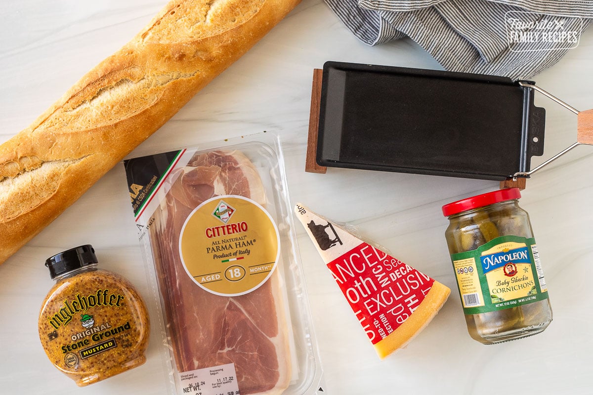 Ingredients to make a raclette sandwich including raclette cheese, French bread, stone, ground, mustard, baby cornichons, prosciutto and a raclette warmer.