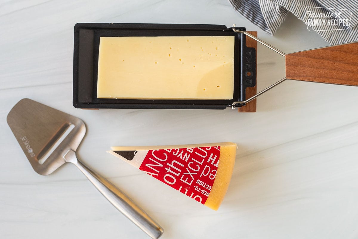 Slice of cheese on a warmer with a cheese, slicer and wedge of raclette cheese.