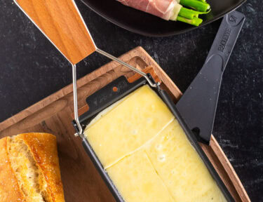 Melted Raclette cheese next to a plate with meat, asparagus, potatoes, pickles, bread and cocktail onions.