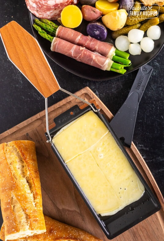 Melted Raclette cheese next to a plate with meat, asparagus, potatoes, pickles, bread and cocktail onions.