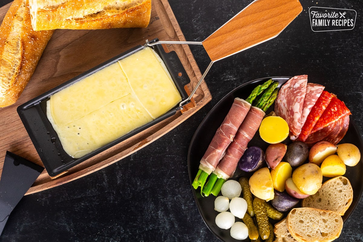 Melted Raclette cheese next to a plate with meat, asparagus, potatoes, pickles, bread and cocktail onions.