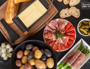 Raclette cheese on board next to plate of meats, small bowl of pickles, baguette bread, asparagus, potatoes and cocktail onions.