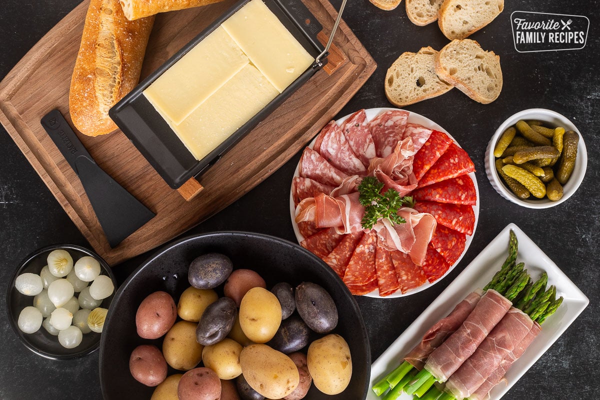 Raclette cheese on board next to plate of meats, small bowl of pickles, baguette bread, asparagus, potatoes and cocktail onions.